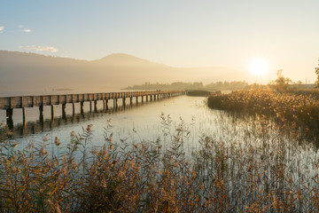 long wooden boardwalk pier over water in golden evening light with a mountain landscape silhouette in the background and golden marsh grass in the foreground