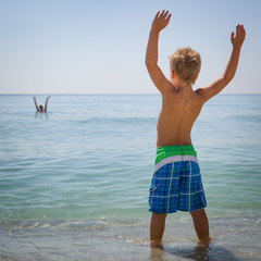 Closeup of a cute kid boy standing by the ocean on a sunny day and waving his arms. The child will swim in the sea. Joy and happiness.