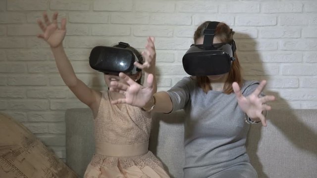 Children play virtual games. Two sisters in virtual reality glasses sit and play at home on the couch against a white brick wall. They wave their hands and look around. Close up. 4K. 25 fps.