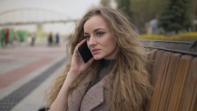 Medium close up beautiful young blonde woman with curly hair in trendy coat phone coversation sitting in the city park on bench on a warm day looking at the camera and smiling
