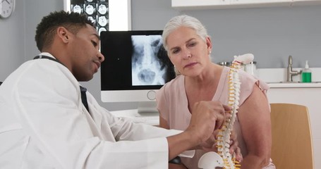 Senior female patient consulting with young doctor about back injury. Close up of black doctor holding model of spinal cord and explaining to elderly woman source of her pain
