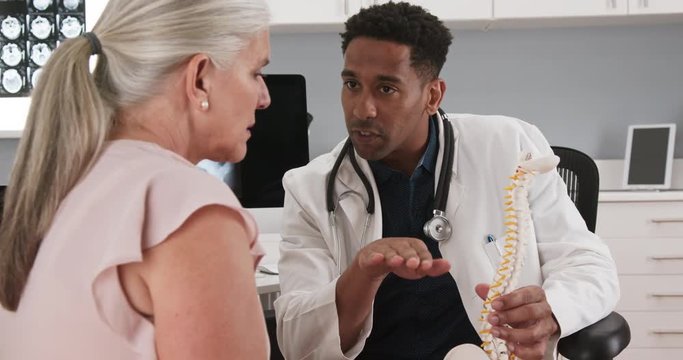 Close up of african-american doctor consulting with elderly patient about spinal cord. Doctor and patient sitting in medical office looking at model of human spine