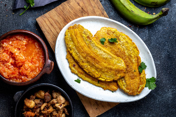 COLOMBIAN CARIBBEAN CENTRAL AMERICAN FOOD. Patacon or toston, fried and flattened whole green plantain banana on white plate with tomato sauce and chicharron Black background, top view