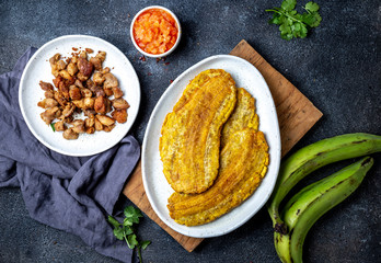 COLOMBIAN CARIBBEAN CENTRAL AMERICAN FOOD. Patacon or toston, fried and flattened whole green plantain banana on white plate with tomato sauce and chicharron Black background, top view