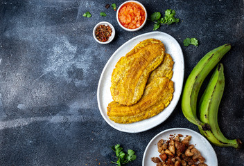 COLOMBIAN CARIBBEAN CENTRAL AMERICAN FOOD. Patacon or toston, fried and flattened whole green plantain banana on white plate with tomato sauce and chicharron Black background, top view