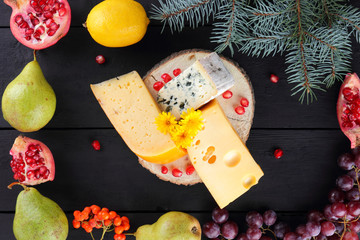 Assortment of cheeses and fruits on dark boards and Christmas tree branches. Swiss cheese, dorblu, Dutch cheese for Christmas. Dairy products, lemon, grapes, pears, pomegranate, rowan berries