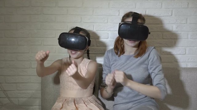 A little girl and her teenage sister in virtual reality glasses sitting and playing virtual games at home on the couch on a white background. They wave their hands and look around. Close up. 4K.