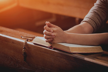 Christian woman praying in church. Hands crossed and Holy Bible on wooden desk.