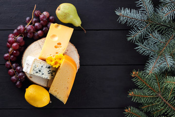 Various types of cheese and fruits on dark boards and Christmas tree branches. Swiss cheese, dorblu, Dutch cheese for Christmas. Dairy products, lemon, grapes, pears. Top view