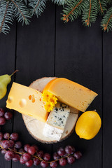 Various types of cheese and fruits on dark boards and Christmas tree branches. Swiss cheese, dorblu, Dutch cheese for Christmas. Dairy products, lemon, grapes, pears. Top view