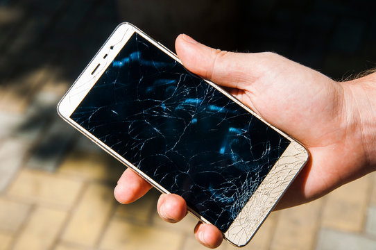 A man's hand holds a mobile phone with a broken screen on the background of the paving stones on a clear day. Broken smartphone. Small shards. Black smashed screen. Unlucky day. Insurance. Watch strap