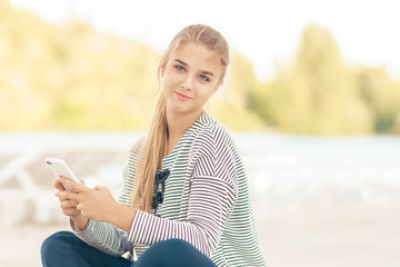 Woman with a smartphone on the beach outside. Vacation and technology. Cute stylish happy girl in casual clothes sitting on a beach near sea using the phone outdoors. Attractive caucasian model