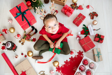 Curious girl wearing xmas costume reindeer antlers sitting on the floor, opening christmas present,...
