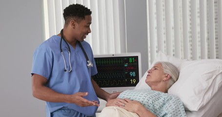 Young black nurse talking to senior woman resting in hospital bed. Male RN assisting elderly female patient lying down in bed - Powered by Adobe