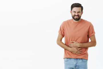 Guy regrets eating fast food feeling pain in stomach clenching teeth and closing eyes from pain touching belly suffering disorder or stomachache waiting for ambulance posing against gray background