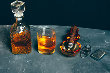 Strong alcoholic drink, scotch whisky in old fashion glass and decanter with smoking cigar in ashtray on gray concrete background