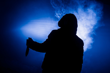 A dangerous hooded man standing in the dark and holding a knife. Face can not be seen. Committing a crime concept. Selective focus