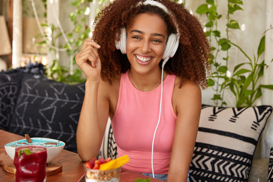 Happy Afro American female meloman listens music with high volume in headphones, sits in cafeteria, eats delicious desserts and exotic dish, connected to modern device, wears casual clothes.