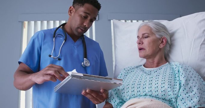 Young male registered nurse reviewing health record with senior female patient. African-American RN looking over doctors notes with elderly woman resting in hospital bed