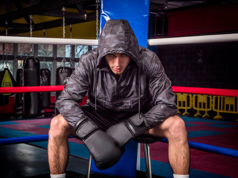 Serious adult man in hoodie and boxing gloves sitting in corner of ring looking violently at camera