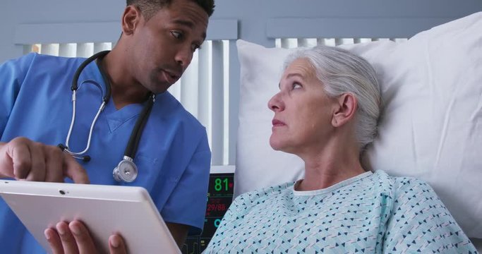 Male millennial black nurse using tablet computer to show senior woman images of her surgery procedure. Young RN and elderly patient looking at electronic pad