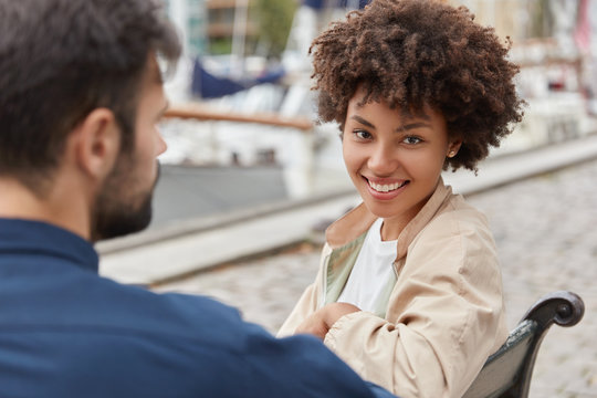 Horizontal shot of cheerful Afro American girl smiles broadly, sits opposite Caucasian unshaven guy, pose at bench, have day off, walk near harbour, enjoy togetherness. People, joy, relationship - Powered by Adobe