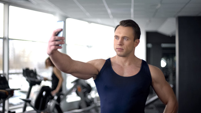 Bodybuilder taking selfie in gym and sending by smartphone, modern technology