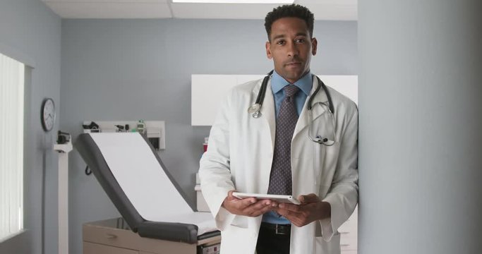 Portrait of handsome male african-american doctor leaned against wall inside medical clinic. Good looking black physician holding tablet computer at doctors office and looking at camera
