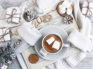 A cup of coffee with marshmallows stands on the open book. A warm sweater. New Year's and Christmas. Christmas tree and gifts of jewelry, bracelets with charms. Huggy concept.