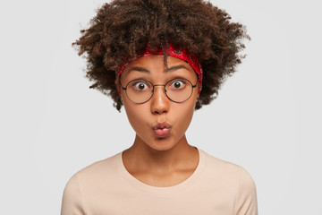 Close up shot of pretty mixed race young woman with curly dark hair, makes grimace at camera, keeps...