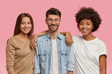 Horizontal shot of three mixed race teenagers spend time together, pose for common photo against pink background. Satisfied guy in eyewear and denim shirt stands between two cheerful women indoor