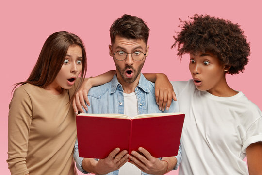 Two mixed females lean at their stylish male groupmate, stand together and stare at book with shocking gaze, read something astonishing, learn information for exam, isolated over pink background