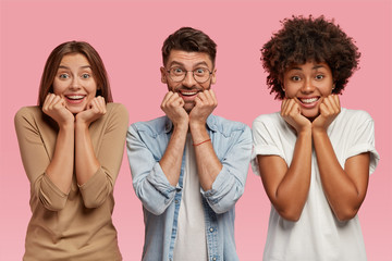 Photo of three multiethnic friends hold chins, watch comedy together during leisure time, wear casual clothes, stand next to each other over pink background. Family portrait. Two sisters and brother