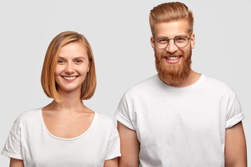 Horizontal shot of cheerful male and female dressed in casual white t shirts, smile gladfully, cooperate together, pose against studio background. Ginger man with thick beard stands near sister