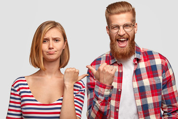 Displeased woman with short hair, frowns face, indicates at ginger guy who smile gladfully, stand...
