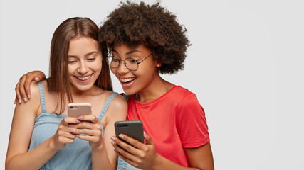 Cheerful mixed race young women watch funny video from social networks, view common photos, have happy facial expressions, hug against white background with free space for your promotion, slogan