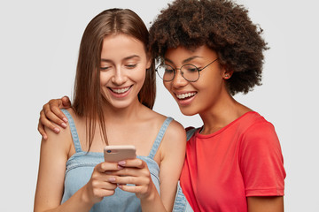 Photo of pretty two multiethnic sisters make booking together on web page via cell phone, watch video online, embrace, isolated over white background. Afro girl has fun with best friend indoor