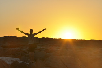 Fototapeta premium Young isolated man's silhouette with outstretched raised hands at sunset, sun rays flare on camera. Traveling and Mindfulness icons
