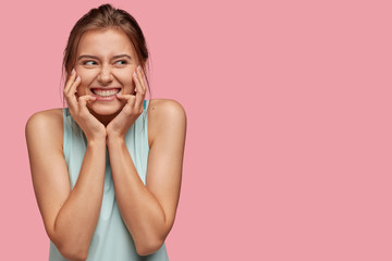 Photo of cheerful content young woman with pleasant smile, keeps hands on face, smiles pleasantly, being pleased by something good, looks happily aside, free space on right side for your slogan