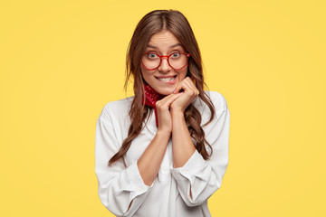 Pretty dark haired woman feels temptation, bites lower lip, looks with great desire, keeps hands near face, wears oversized white shirt, models against yellow background, has red rimed spectacles