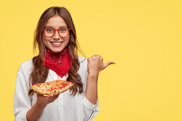 Glad smiling woman holds tasty pizza, indicates with thumb aside as shows place where she bought it, advertises pizzeria, wears red bandana and white shirt, isolated over yellow studio wall.