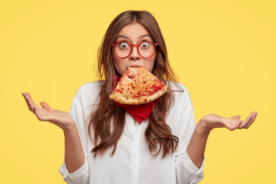 Surprised Caucasian woman keeps slice of pizza in mouth, spreads hands with hesitation, has puzzled facial expression, dressed in white shirt, eats junk food, models against yellow background