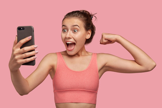 Happy girl with healthy skin, has fun, makes video call, shows musles in camera, wears casual pink top, feels proud of herself. Caucasian lady takes selfie, models indoor. People, sport, technology