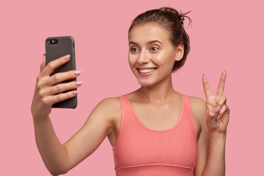 Studio shot of glad female with toothy smile, combed hair, has sporty body, makes peace sign or V gesture at camera of cell phone, poses for making selfie, isolated over pink background. Video call