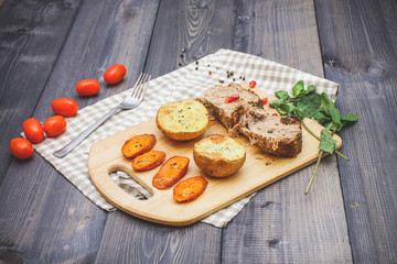 On a cutting board, slices of baked meat, baked potatoes and carrots, linen checkered napkin and cherry tomatoes on a dark graphite surface