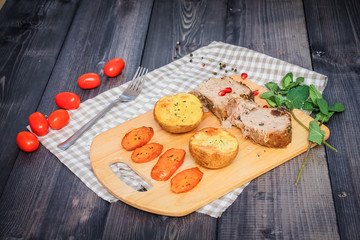 On a cutting board, slices of baked meat, baked potatoes and carrots, linen checkered napkin and cherry tomatoes on a dark graphite surface