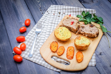 On a cutting board, slices of baked meat, baked potatoes and carrots, linen checkered napkin and cherry tomatoes on a dark graphite surface