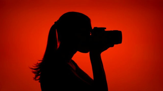 Silhouette of young woman taking photos on professional camera. Female's face in profile taking pictures on red background. Black contour shadow of teenager's half-face photographing