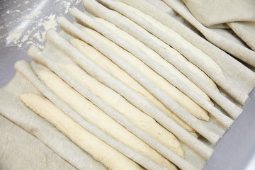 Homemade, imperfectly shaped rustic French baguette breads after final rise waiting to go in the oven for baking with dough cutter and recipe book on floured wooden kitchen table