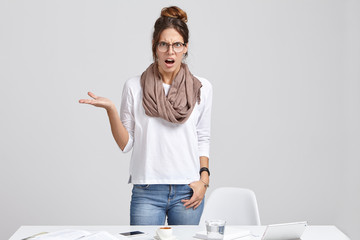 Outraged grumpy woman director gestures, has unhappy annoyed expression, being angry with much work, wears stylish white sweater and jeans, stands at workplace with modern gadgets and coffee